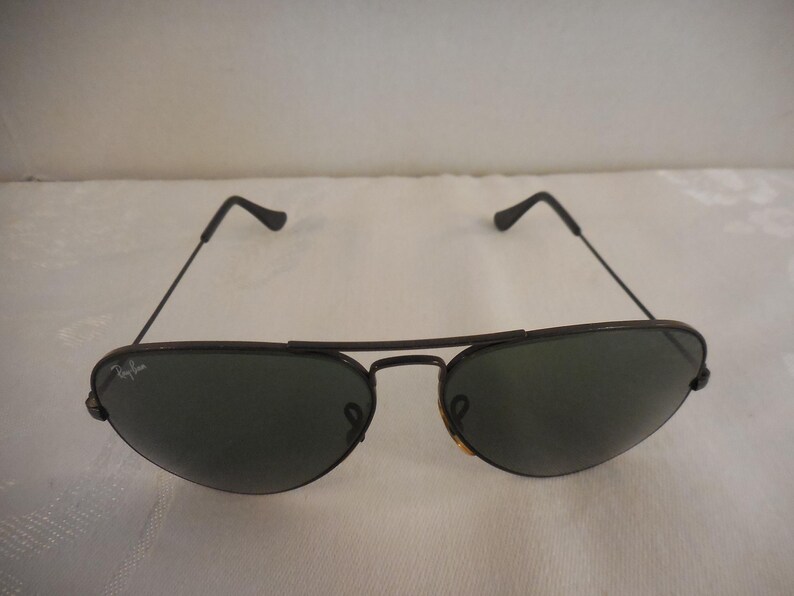 May include: Black aviator sunglasses with dark green lenses. The sunglasses have a thin, dark metal frame and a double bridge. The Ray-Ban logo is visible on the left lens. The sunglasses are a classic style.