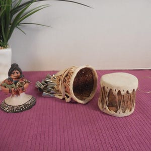 May include: A ceramic figurine of a person holding two small figures, a woven basket containing several silver-colored metal cones, and a small cylindrical drum with a white skin top and laced sides, all arranged on a textured surface.