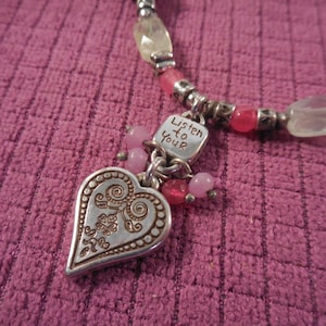May include: A silver-toned necklace with a heart pendant and the words "Listen to Your" engraved on a small square charm. The necklace features pink and clear beads, with a textured pink background.