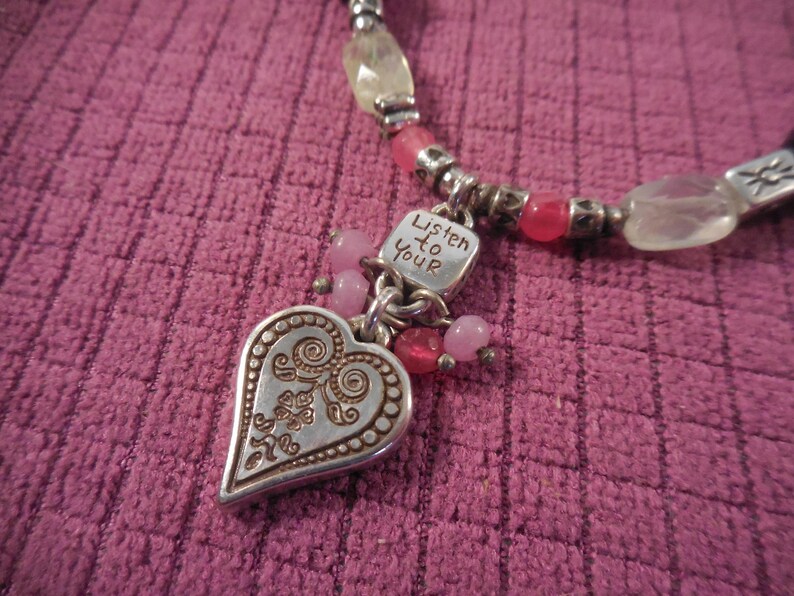 May include: A silver-toned necklace with a heart pendant and the words "Listen to Your" engraved on a small square charm. The necklace features pink and clear beads, with a textured pink background.