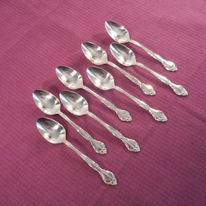 May include: Eight silver spoons with ornate handles are arranged diagonally on a deep purple surface. The spoons have a reflective, polished finish, and the handles feature a detailed floral design. The spoons are likely for serving or dessert.