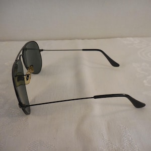 May include: Black aviator sunglasses with dark lenses and a thin, black metal frame. The sunglasses have a classic teardrop shape and gold-colored accents near the nose bridge. The temples curve down at the ends.