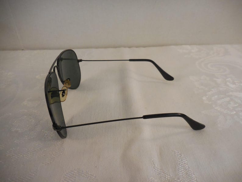 May include: Black aviator sunglasses with dark lenses and a thin, black metal frame. The sunglasses have a classic teardrop shape and gold-colored accents near the nose bridge. The temples curve down at the ends.