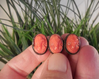 May include: A ring featuring three oval-shaped, salmon-colored coral cameos set in a dark metal band. Each cameo displays a carved profile of a face. The metal setting has a textured, decorative edge surrounding each individual coral piece.