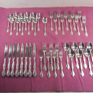 May include: A complete set of silver-colored flatware, including spoons, forks, and knives, arranged on a pink tablecloth. The silverware has decorative handles and is neatly organized, ready for a formal dining setting.