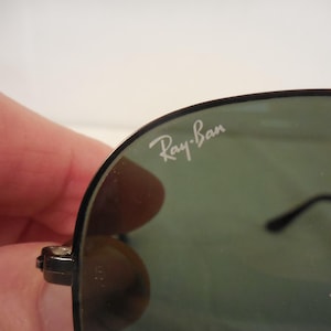 May include: Close-up of Ray-Ban sunglasses. The sunglasses have a black frame and dark green lenses. The Ray-Ban logo is visible in white on the upper left lens. The sunglasses are held by a person.