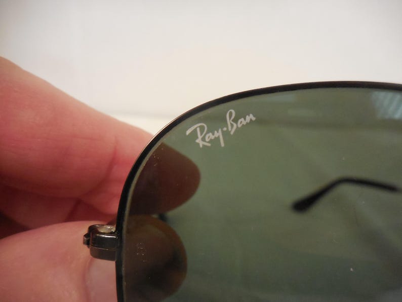 May include: Close-up of Ray-Ban sunglasses. The sunglasses have a black frame and dark green lenses. The Ray-Ban logo is visible in white on the upper left lens. The sunglasses are held by a person.