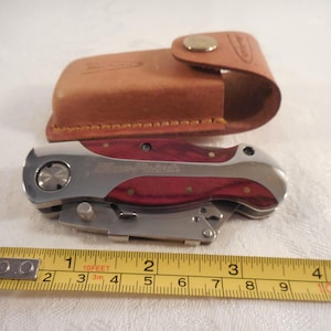 May include: A folding utility knife with a wood grain handle and silver metal accents, featuring the text Blue-Point. The knife is positioned next to a yellow measuring tape showing a length of approximately 4 inches, accompanied by a tan leather sheath.