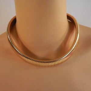 May include: A gold-colored, ribbed collar necklace. The necklace is thick and flexible, designed to sit close to the neck. The necklace is displayed on a mannequin.