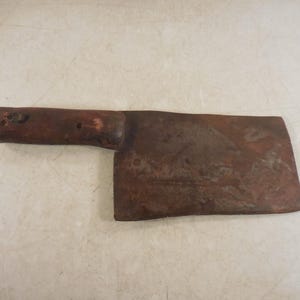 May include: A vintage cleaver with a dark wooden handle and a large, rectangular, rusty blade. The blade shows signs of age and wear, with a dark brown color. The handle is a lighter brown and appears to be made of wood.
