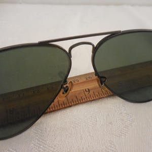 May include: Aviator sunglasses with dark green lenses and a dark metal frame. The sunglasses are resting on a wooden ruler, with the number 19 visible. The Ray-Ban logo is visible on the lens.