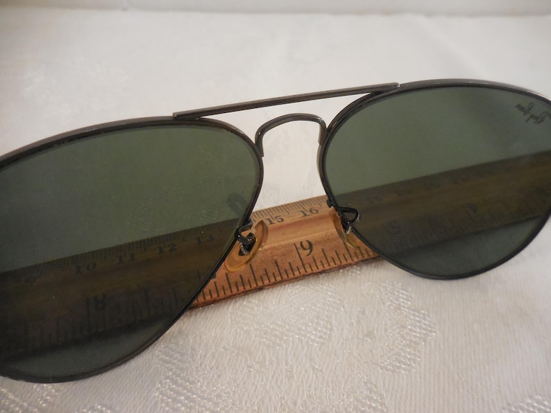 May include: Aviator sunglasses with dark green lenses and a dark metal frame. The sunglasses are resting on a wooden ruler, with the number 19 visible. The Ray-Ban logo is visible on the lens.