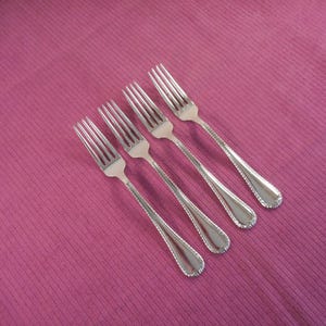 May include: Four silver forks with a decorative handle design are arranged diagonally on a pink textured surface. The forks have tines for eating and a detailed, textured handle. The background is a solid pink color.