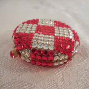 May include: A round, bejeweled trinket box with a red and white checkerboard pattern. The box is covered in sparkling red and clear rhinestones, with a gold-colored metal clasp.