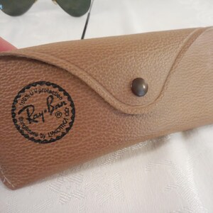 May include: Tan leather sunglasses case with a snap closure. The case features the Ray-Ban logo in a circular design. The case is designed to protect sunglasses and has a textured surface.