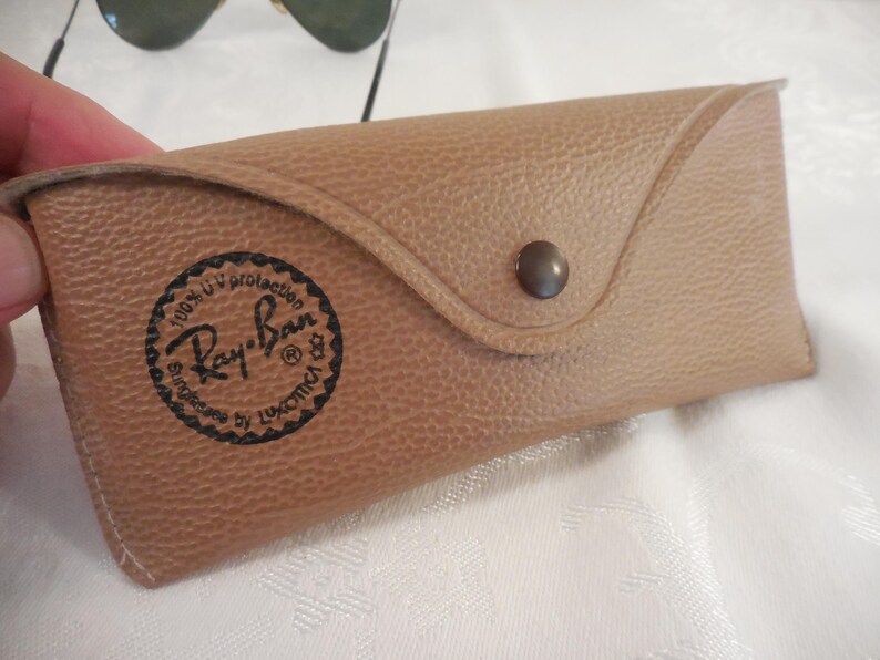 May include: Tan leather sunglasses case with a snap closure. The case features the Ray-Ban logo in a circular design. The case is designed to protect sunglasses and has a textured surface.