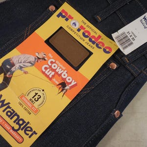 May include: A pair of blue denim jeans with a Wrangler label and a Rodeo competition jeans tag. The tag features a cowboy riding a horse and the text "The Original Cowboy Cut Jeans".