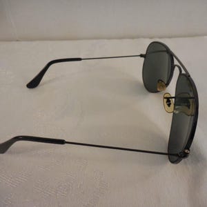 May include: Black aviator sunglasses with dark lenses. The sunglasses have a thin, black metal frame and temples. The lenses are oval-shaped and the nose pads are a light tan color. The sunglasses are resting on a white surface.