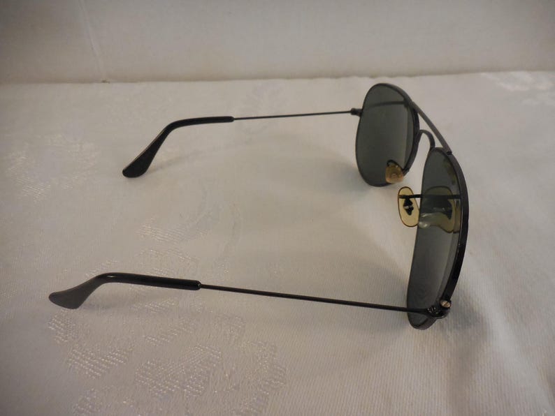 May include: Black aviator sunglasses with dark lenses. The sunglasses have a thin, black metal frame and temples. The lenses are oval-shaped and the nose pads are a light tan color. The sunglasses are resting on a white surface.