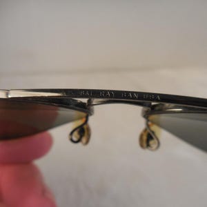 May include: Close-up of Ray-Ban sunglasses. The silver frames have dark lenses and the text "RAY BAN USA" is visible on the frame. The sunglasses are held by a person.