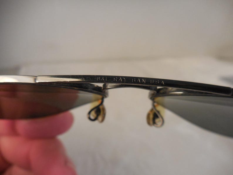 May include: Close-up of Ray-Ban sunglasses. The silver frames have dark lenses and the text "RAY BAN USA" is visible on the frame. The sunglasses are held by a person.
