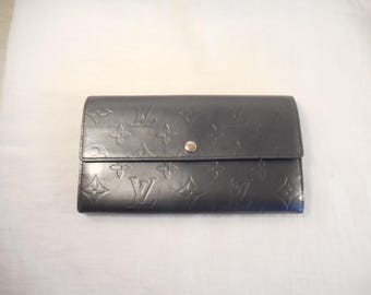 May include: A dark gray leather wallet with embossed floral and logo patterns. The wallet has a snap closure and a rectangular shape. The design includes the brand's signature monogram. The wallet is likely designed for carrying cash and cards.