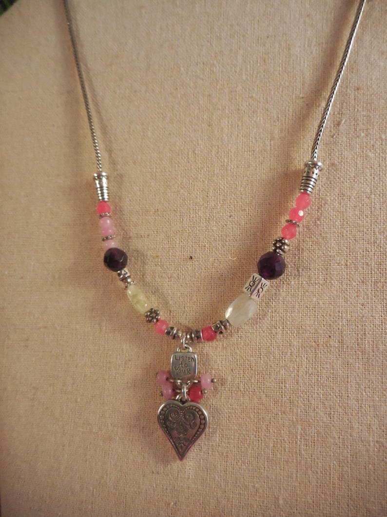 May include: A silver necklace with a heart pendant and colorful beads. The pendant has a heart shape with an ornate design and the words "Listen to Your Heart". The beads are pink, purple, and white.