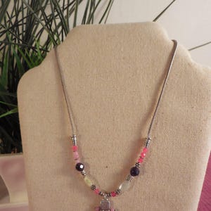 May include: A silver necklace with a beaded design. The necklace features a variety of beads in pink, purple, and white, with a heart-shaped pendant. The necklace is displayed on a beige bust.