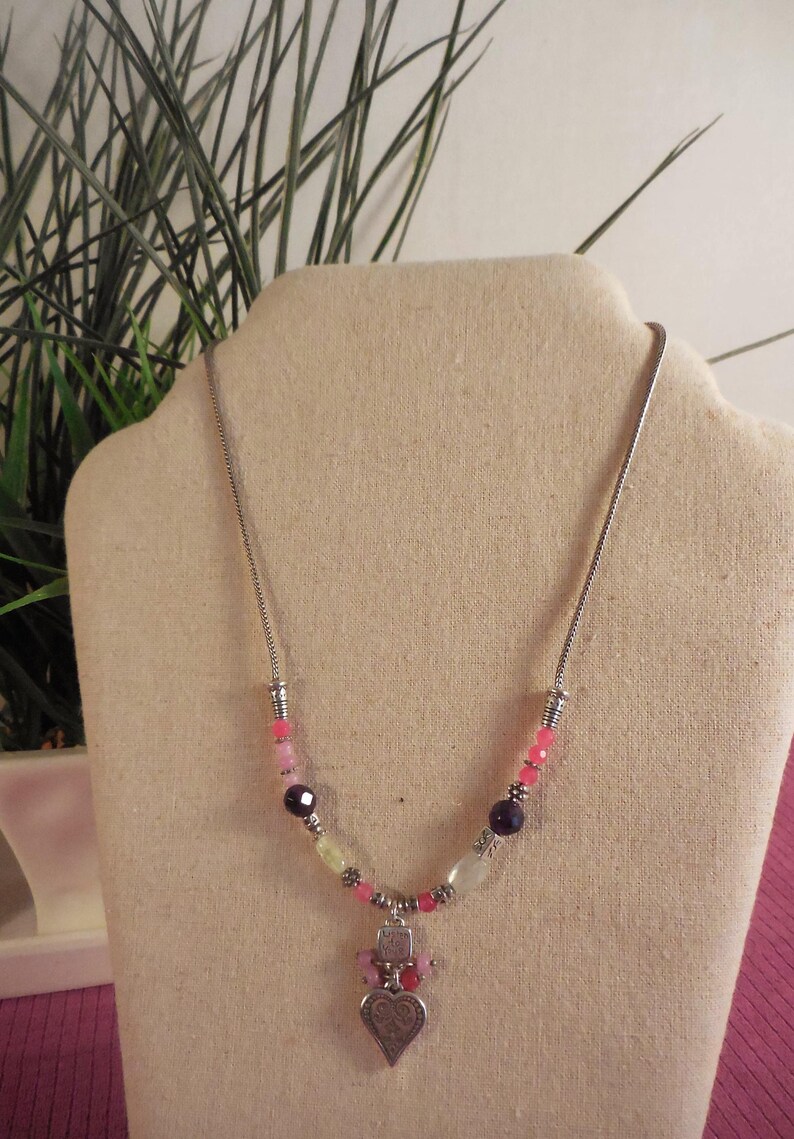 May include: A silver necklace with a beaded design. The necklace features a variety of beads in pink, purple, and white, with a heart-shaped pendant. The necklace is displayed on a beige bust.