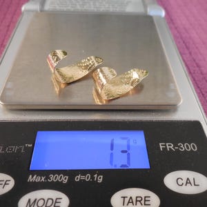May include: Two gold-colored, curved earrings with textured surfaces are displayed on a digital scale. The scale shows a weight of 1.3 grams. The scale has a blue digital display and black buttons.