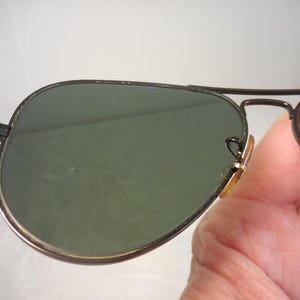 May include: Aviator-style sunglasses with a black metal frame and green lenses. The lenses have a slight gradient, and the frame features a double bridge design. The sunglasses are held by a person.