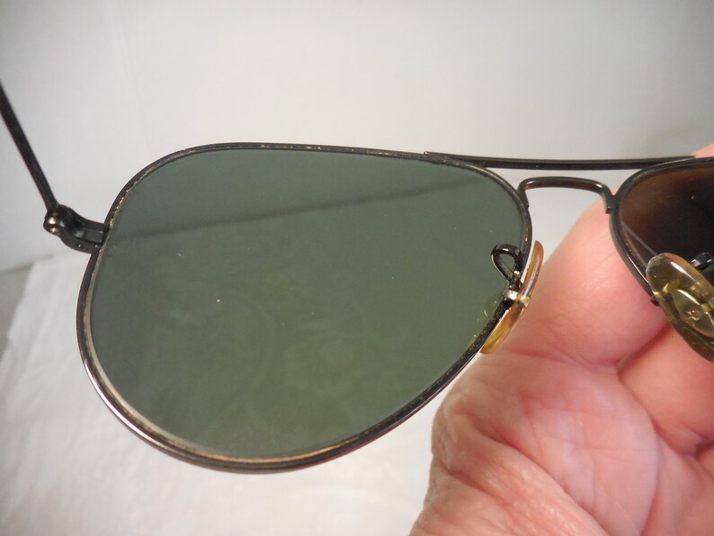 May include: Aviator-style sunglasses with a black metal frame and green lenses. The lenses have a slight gradient, and the frame features a double bridge design. The sunglasses are held by a person.