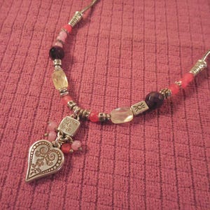 May include: A silver necklace with a heart pendant and colorful beads. The heart pendant has an ornate design and the words "Listen to Your" engraved on a small square charm. The beads are pink, red, yellow, and dark purple.