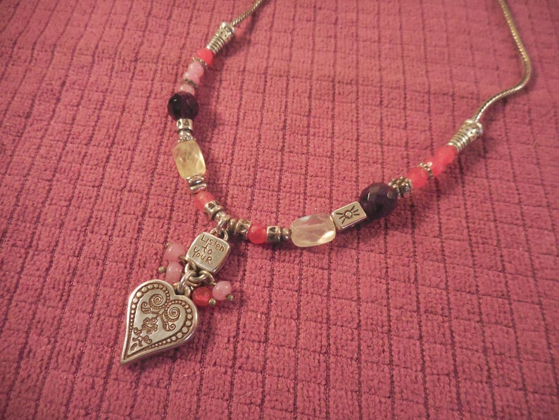 May include: A silver necklace with a heart pendant and colorful beads. The heart pendant has an ornate design and the words "Listen to Your" engraved on a small square charm. The beads are pink, red, yellow, and dark purple.