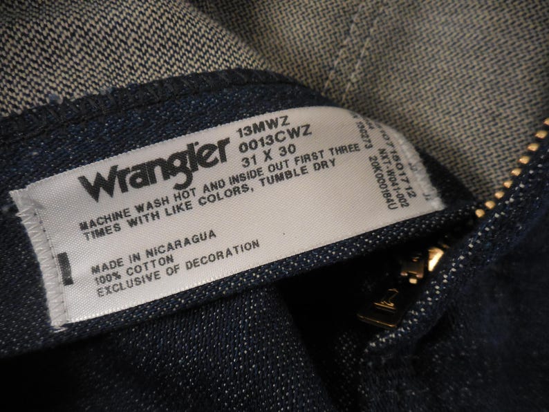 May include: A white Wrangler jeans care label with black text. The label says "Machine wash hot and inside out first three times with like colors, tumble dry. Made in Nicaragua, 100% cotton, exclusive of decoration. 13MWZ 0013Cwz 31 x 30."