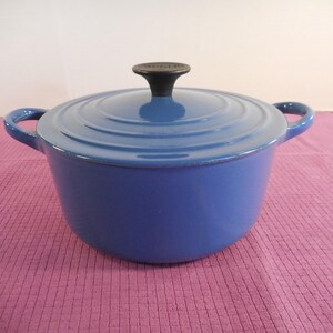May include: A blue cast iron Dutch oven with a lid and two side handles. The lid has a black knob. The pot is a vibrant blue color and sits on a pink surface. A kitchenware item.