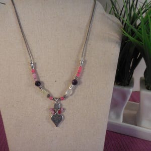 May include: A silver-toned necklace with a heart pendant. The necklace features a variety of beads in pink, purple, and clear colors. The heart pendant is detailed with a textured surface. The necklace is displayed on a mannequin.