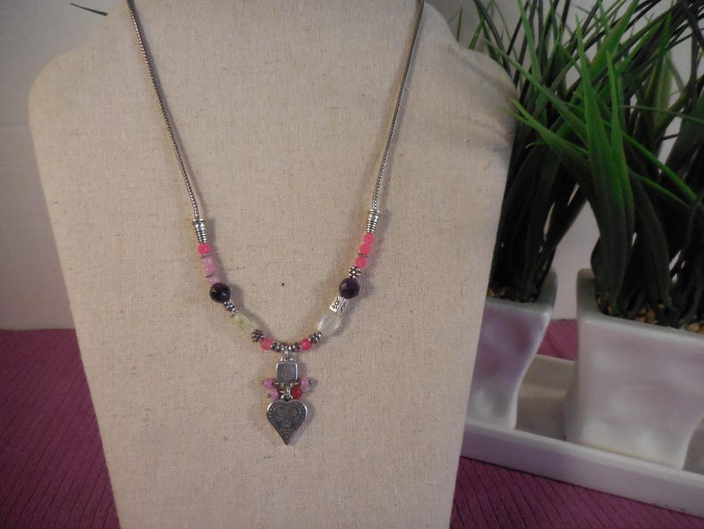 May include: A silver-toned necklace with a heart pendant. The necklace features a variety of beads in pink, purple, and clear colors. The heart pendant is detailed with a textured surface. The necklace is displayed on a mannequin.