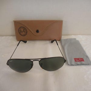 May include: Black aviator sunglasses with green lenses, a brown leather case, and a gray cleaning cloth. The sunglasses have a thin black frame and the Ray-Ban logo is visible on the case and cloth. The case has a snap closure.