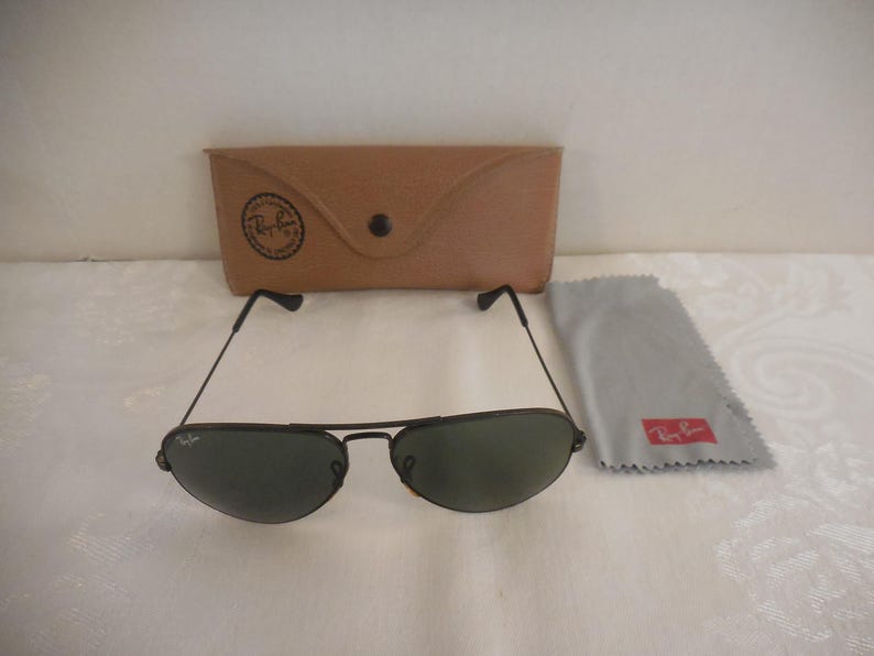May include: Black aviator sunglasses with green lenses, a brown leather case, and a gray cleaning cloth. The sunglasses have a thin black frame and the Ray-Ban logo is visible on the case and cloth. The case has a snap closure.