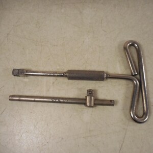 May include: Three metal tools are displayed on a light-colored surface. One is a T-handle wrench, another is a socket wrench with a knurled handle, and the third is a sliding T-handle. These tools are likely used for mechanical or automotive work.