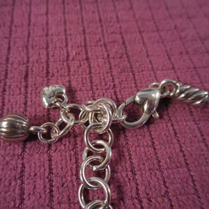 May include: Close-up of a silver-toned necklace clasp and chain. The clasp is connected to a chain of small rings and a decorative charm. The background is a textured, dark pink fabric.