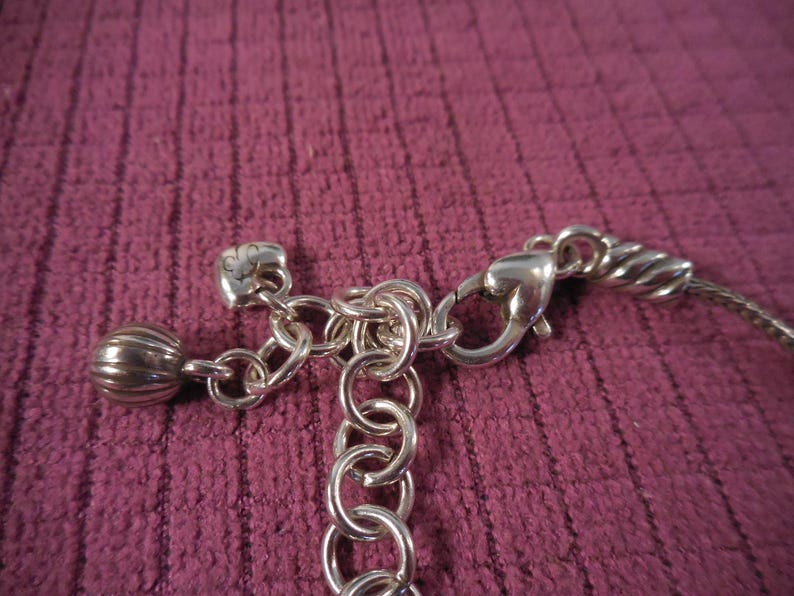 May include: Close-up of a silver-toned necklace clasp and chain. The clasp is connected to a chain of small rings and a decorative charm. The background is a textured, dark pink fabric.
