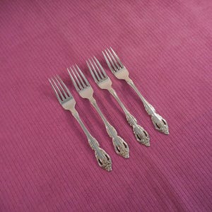 May include: Four silver forks with ornate handles are arranged diagonally on a textured, magenta surface. The forks have three tines and decorative detailing on the handles, suggesting a vintage or antique style. The overall composition is simple and elegant.