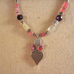 May include: A necklace with a silver heart pendant and the words "Listen to Your" engraved on a small tag. The necklace features pink, purple, and white beads. The heart pendant has an ornate design.