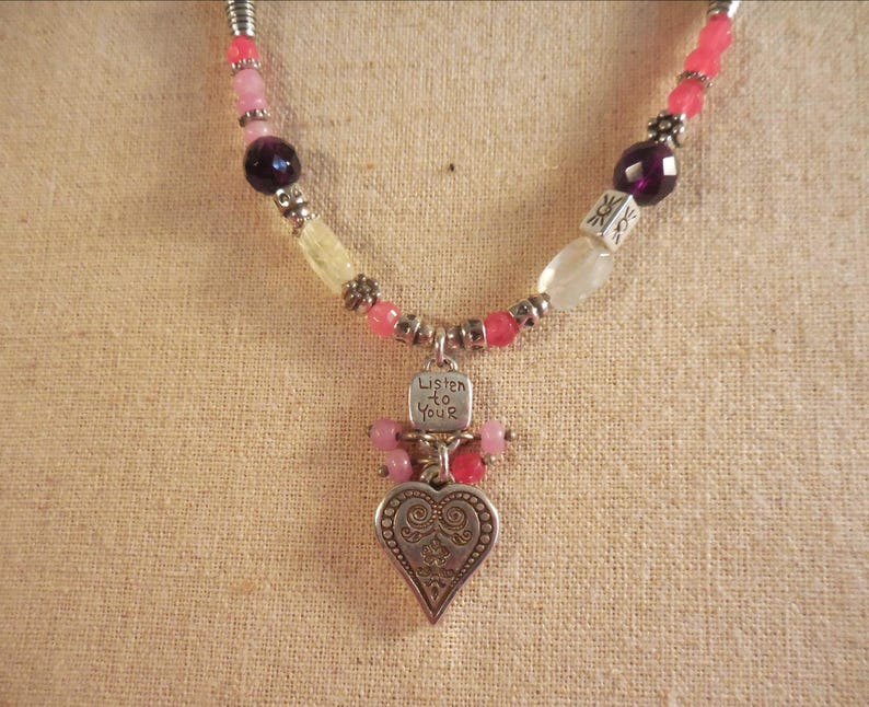 May include: A necklace with a silver heart pendant and the words "Listen to Your" engraved on a small tag. The necklace features pink, purple, and white beads. The heart pendant has an ornate design.