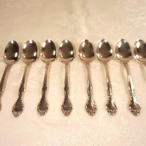 May include: A set of eight silver spoons arranged in a row. Each spoon has a rounded bowl and a decorative handle with an ornate floral design. The spoons are displayed on a white, textured surface.