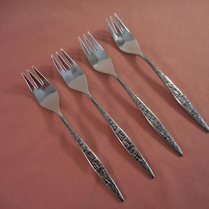 May include: Four silver forks with a textured handle design are arranged diagonally on a reddish-brown surface. The forks have three tines and a polished finish, reflecting light. The handles feature a geometric pattern.