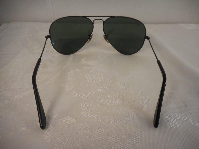 May include: Black aviator sunglasses with dark green lenses. The sunglasses have a thin, black metal frame and temples. The lenses are teardrop-shaped. The bridge and hinges are gold-colored. A classic style for eye protection.