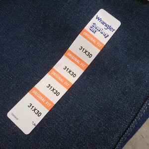 May include: A close-up of a blue denim fabric with a white tag that reads "Wrangler Cowboy Cut Original Fit" and lists the size "31X30" four times.